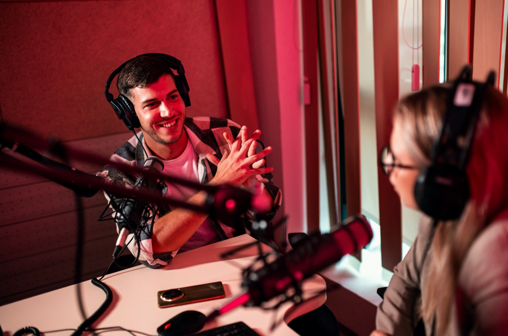 Man and woman in red-lit booth chatting enthusiastically into mounted microphones.