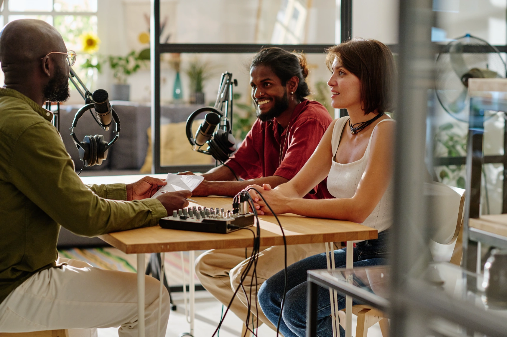 Three podcasters at a wooden table with laptops and mics in a bright home setting.