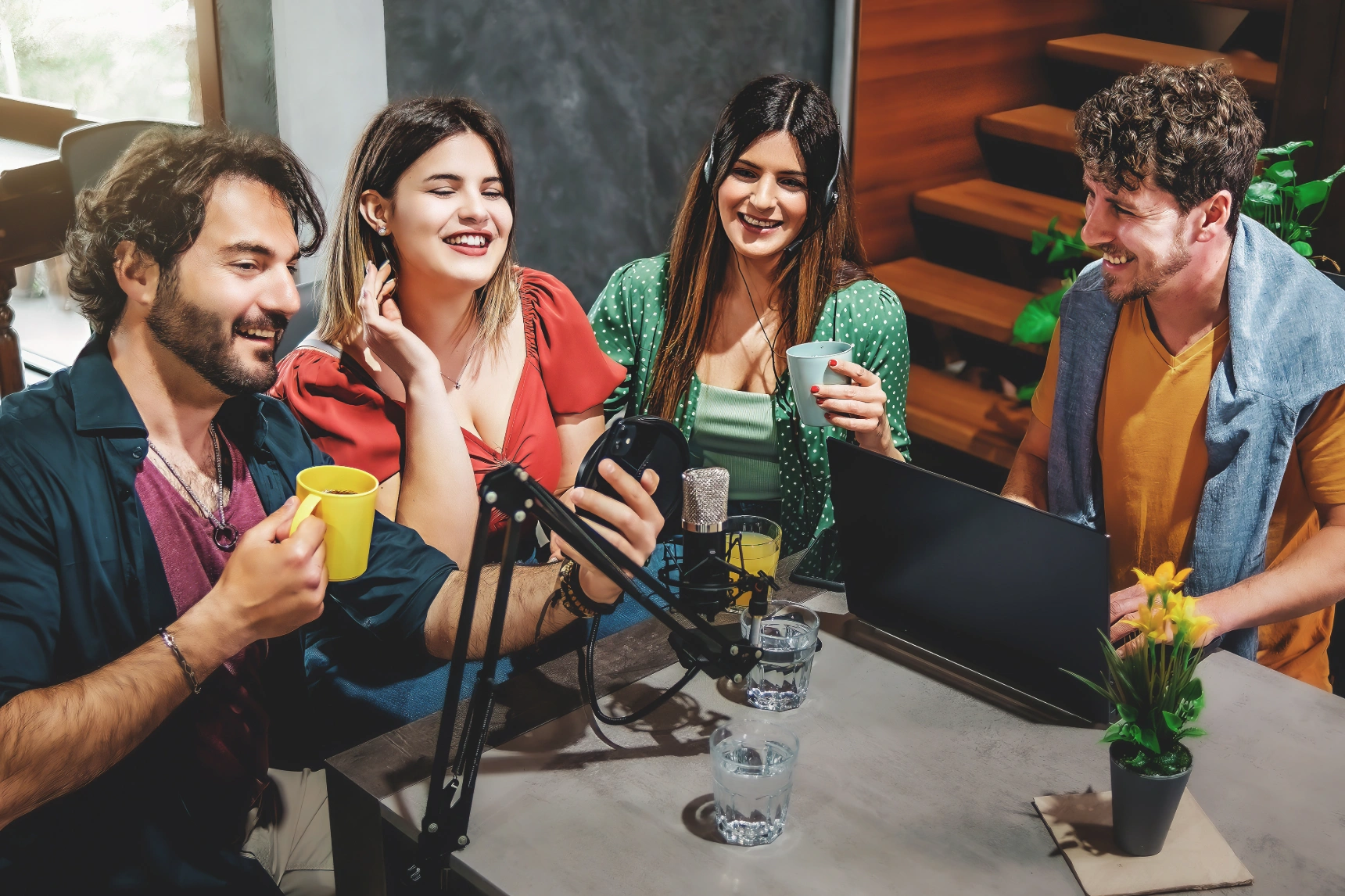 Four friends around a table recording a casual podcast while holding drinks.