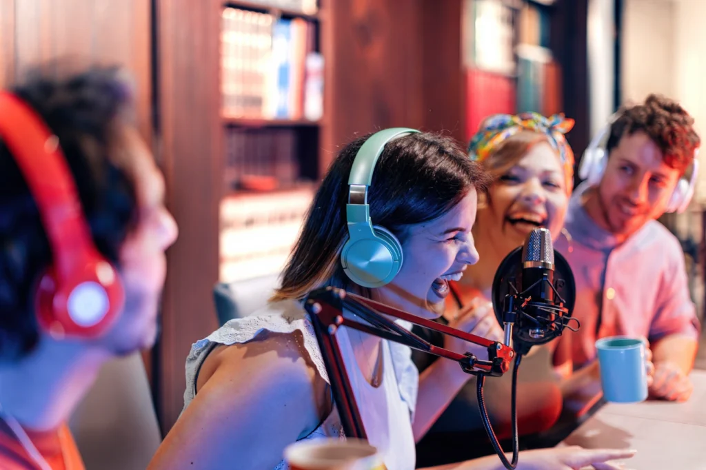 Woman in teal headphones laughs into studio mic while friends cheer behind her.