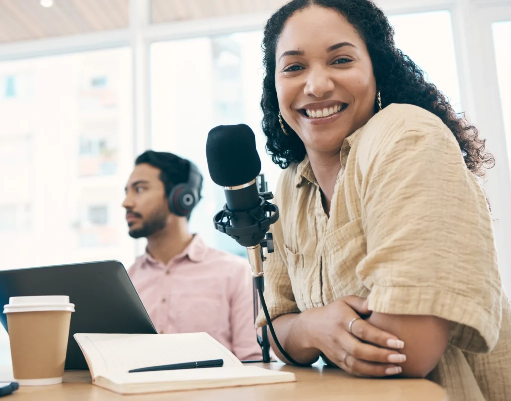 Smiling female host leans by microphone with notebook while male co-host uses laptop.