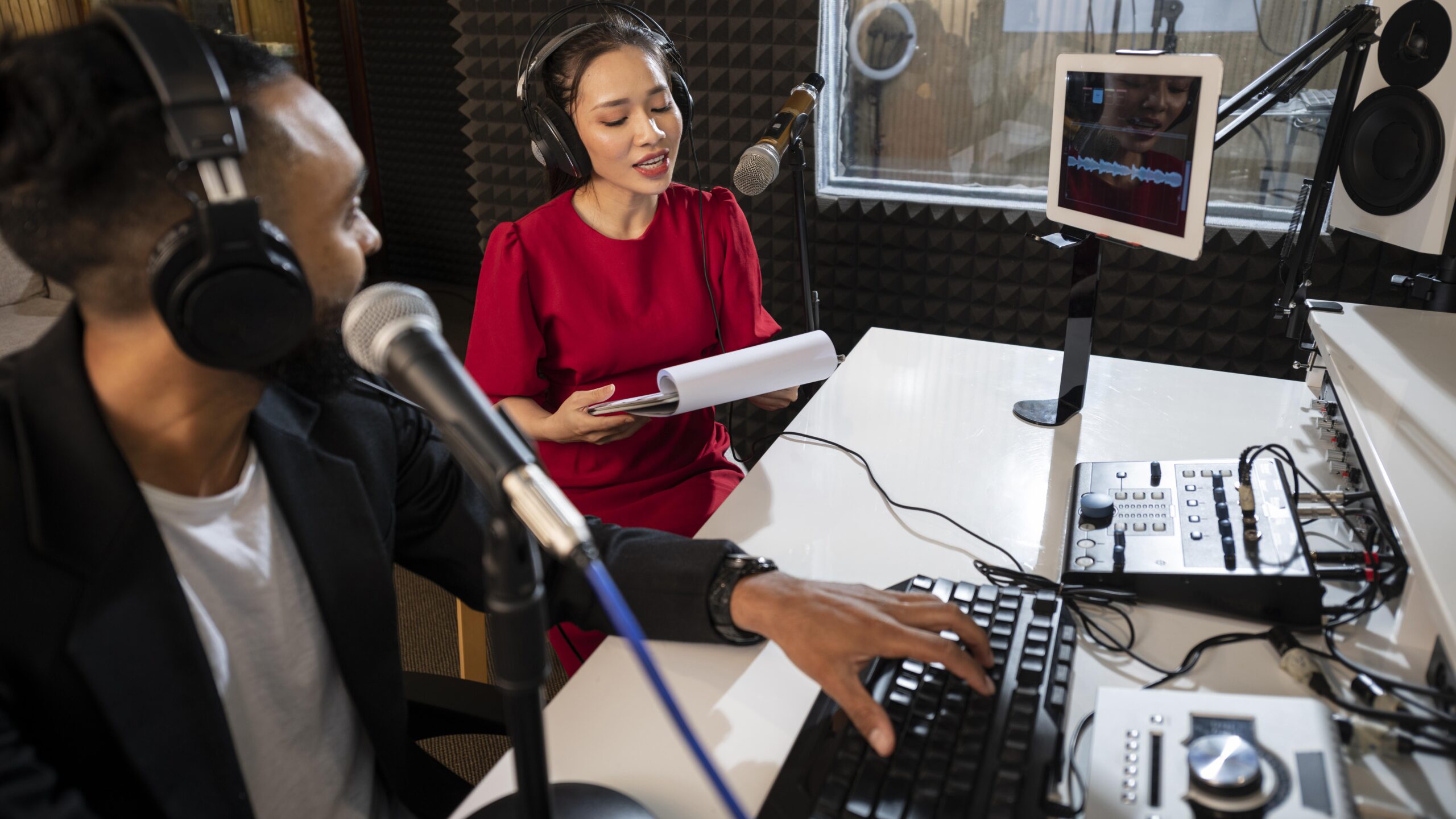 Two hosts in an acoustically treated booth recording while one types at a keyboard.