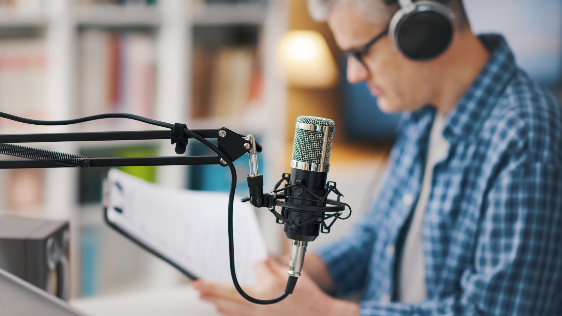Image of a person recording a podcast, wearing headphones and reading from a script, with a professional microphone in focus against a blurred background.