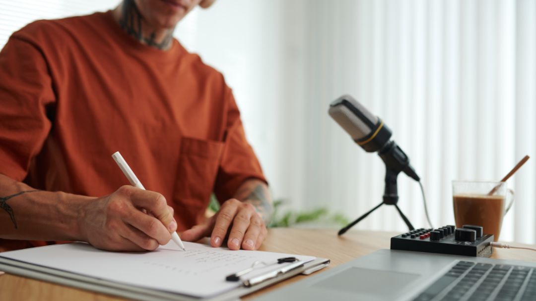 Image for How to Start a Podcast Script, showing a person with tattoos writing on a clipboard next to a microphone, laptop, and small mixer.