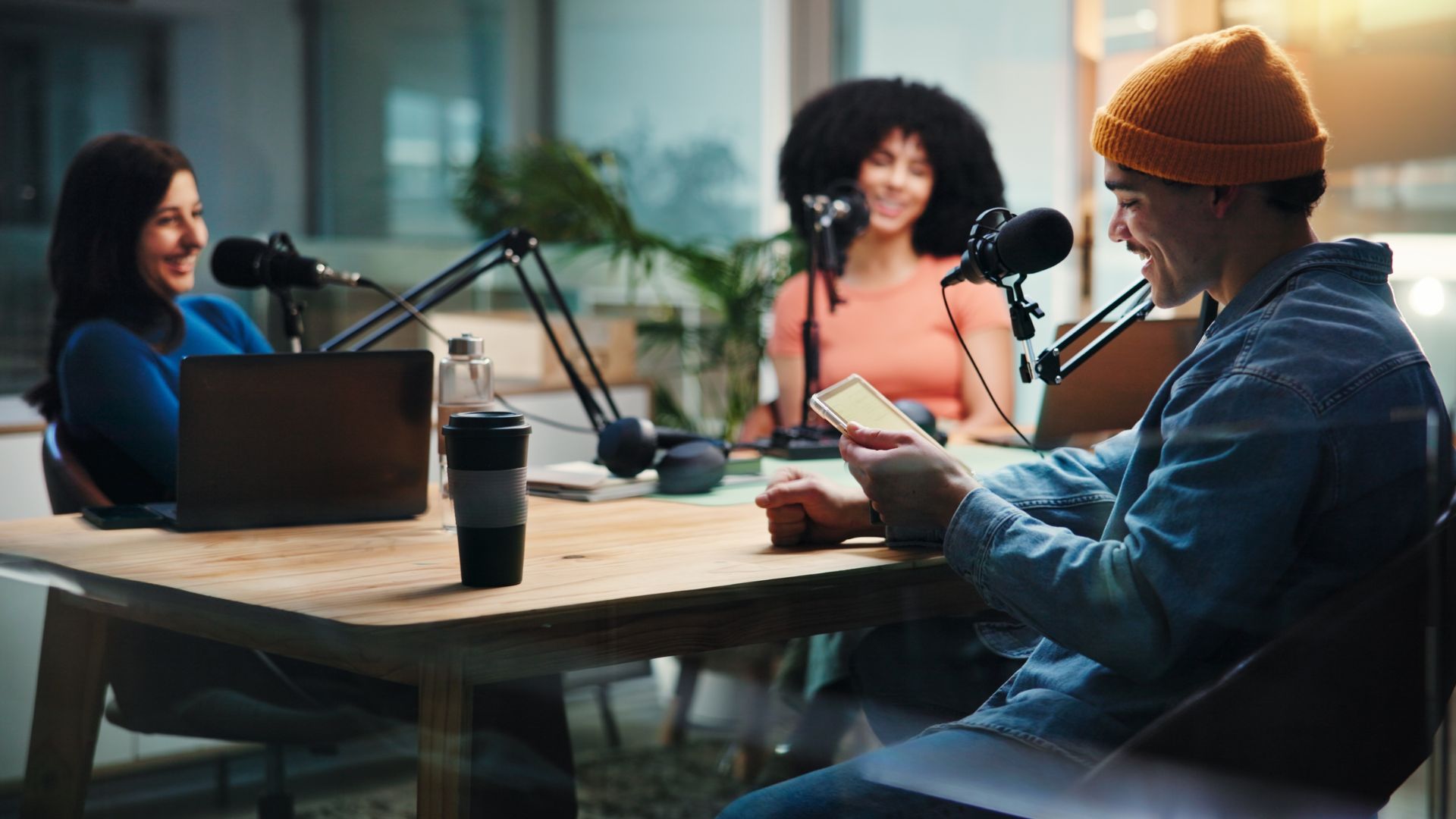 Three diverse people sitting around a table with professional microphones, recording a podcast episode, with one person smiling while checking their phone.