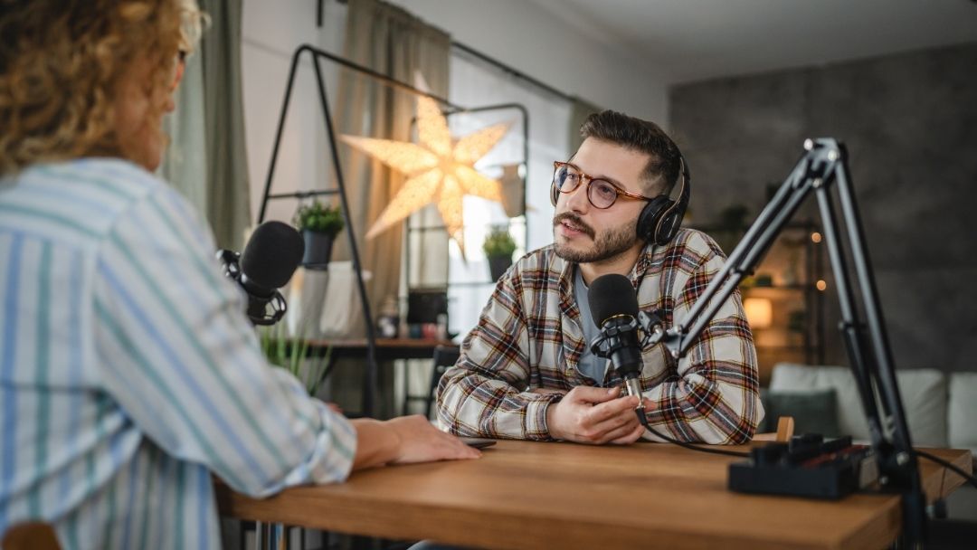 Podcast host with headphones interviewing guest at microphones in home studio, demonstrating how to find podcast guests in 2026 that say yes and show up.