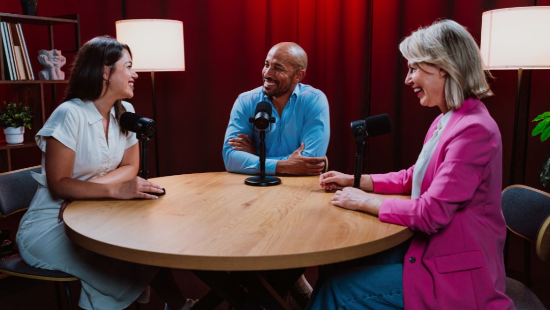Internal vs External Podcast Host Which One Works Best for Your Brand. Three people laughing around a wooden table with microphones during a lively podcast recording session: woman in white dress, man in blue shirt, and woman in pink blazer.