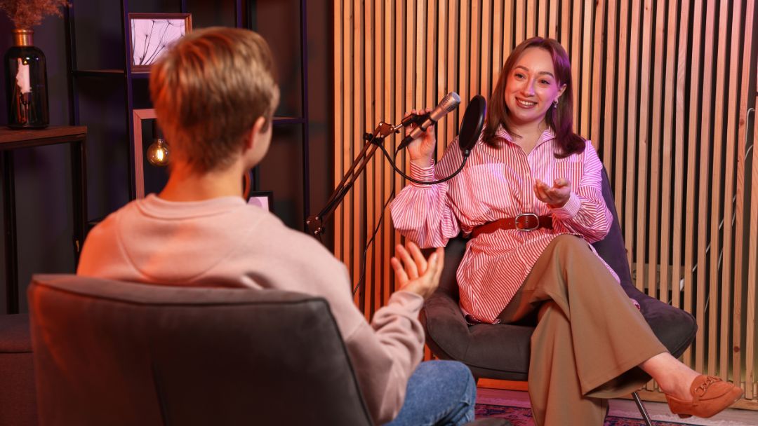 Podcast recording session: Smiling woman in striped shirt speaking into a professional microphone while gesturing to a man in a hoodie, during an interview in a modern studio setup.